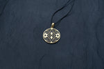 A round Shipibo pendant from Kaya Movement's Shamanic jewelry collection rests on a thin black cord against a dark textured background. This intricate Icaros pendant showcases a detailed geometric design in oxidized yellow brass and black, featuring symmetrical patterns with small cross shapes and other decorative elements.