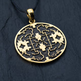 A gold, round Kaya Movement Shipibo Icaro pendant hangs from a black cord against a textured dark background. The design features symmetrical geometric and maze-like shapes within the circular frame, accented with beautifully oxidized details.