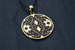 A gold, round Kaya Movement Shipibo Icaro pendant hangs from a black cord against a textured dark background. The design features symmetrical geometric and maze-like shapes within the circular frame, accented with beautifully oxidized details.