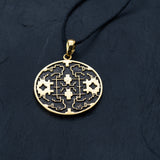 A round gold Shipibo Icaros Pendant from Kaya Movement, showcasing intricate symmetrical, maze-like structures and floral motifs with oxidized details and a hollow center, hangs from a thin black cord. This psychedelic and shamanic piece of jewelry is displayed on a dark, textured surface, highlighting its exceptional craftsmanship.
