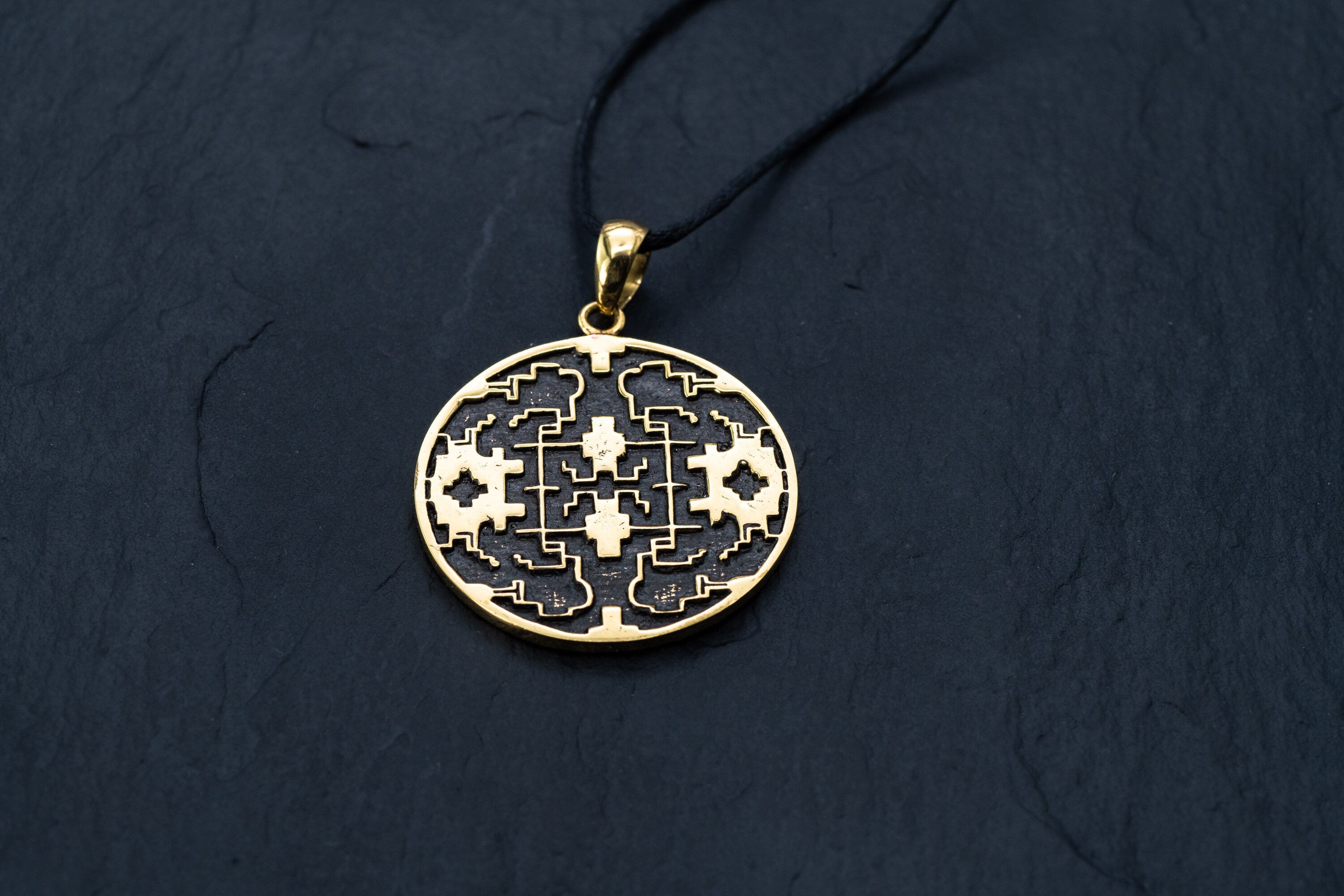 A round gold Shipibo Icaros Pendant from Kaya Movement, showcasing intricate symmetrical, maze-like structures and floral motifs with oxidized details and a hollow center, hangs from a thin black cord. This psychedelic and shamanic piece of jewelry is displayed on a dark, textured surface, highlighting its exceptional craftsmanship.