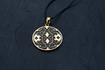 A round gold Shipibo Icaros Pendant from Kaya Movement, showcasing intricate symmetrical, maze-like structures and floral motifs with oxidized details and a hollow center, hangs from a thin black cord. This psychedelic and shamanic piece of jewelry is displayed on a dark, textured surface, highlighting its exceptional craftsmanship.