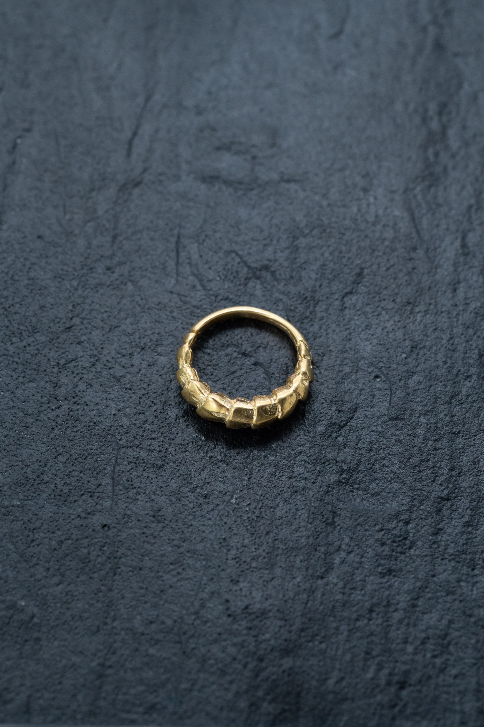A ring from Kaya Movement's collection, the "Biomechanical - 18k Gold Septum 10mm - Alien Xenomorph," lies on a dark, textured surface. Featuring a braided or interwoven pattern around its band, this distinctive and elegant piece resembles cybernetic bio-organic jewelry.