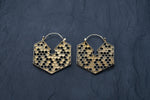 A pair of Kaya Movement's Oversized Hoop Honeycomb Hexagon Bee Gold Brass Earrings, Sacred Geometry Cool Boho Hypercube Jewelry, showcased against a dark textured background. These closed hoop style earrings intricately feature small hexagonal shapes that mimic a beehive structure.