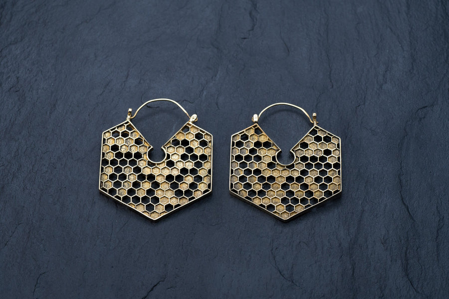 A pair of Kaya Movement's Oversized Hoop Honeycomb Hexagon Bee Gold Brass Earrings, Sacred Geometry Cool Boho Hypercube Jewelry, showcased against a dark textured background. These closed hoop style earrings intricately feature small hexagonal shapes that mimic a beehive structure.