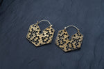 A pair of elegant Kaya Movement Oversized Hoop Honeycomb Hexagon Bee Gold Brass Earrings lay on a dark textured surface. Featuring an intricate honeycomb pattern with a geometric shape, they exude modern sophistication in a closed hoop design, embodying the essence of sacred geometry and cool boho style.