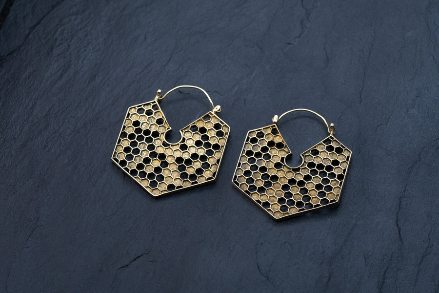 A pair of elegant Kaya Movement Oversized Hoop Honeycomb Hexagon Bee Gold Brass Earrings lay on a dark textured surface. Featuring an intricate honeycomb pattern with a geometric shape, they exude modern sophistication in a closed hoop design, embodying the essence of sacred geometry and cool boho style.