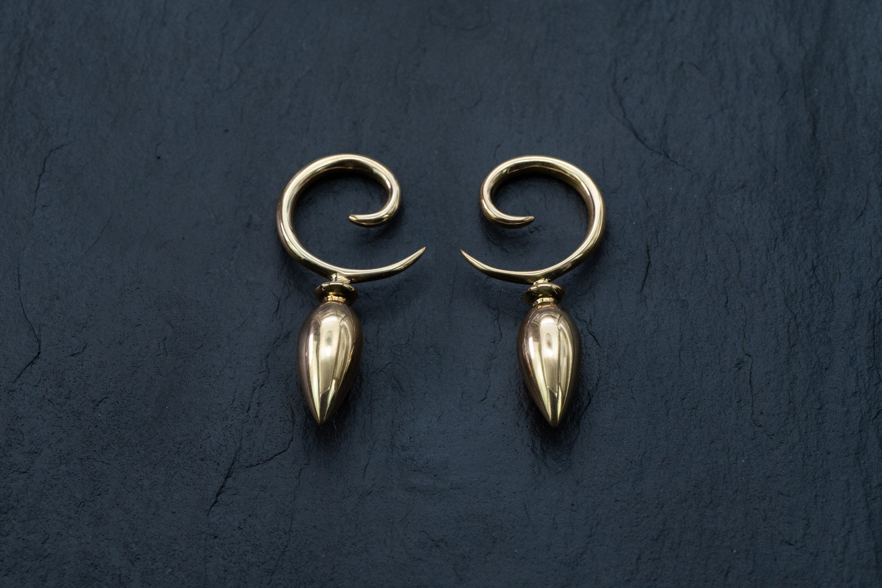 A pair of Drop Ear Hangers - Spiral Gauges by Kaya Movement, crafted as elegant ear weights for stretched ears, are positioned against a dark, textured background. The spirals curve gracefully, and the smooth, polished gold surface of the 4g ear weights reflects light subtly.