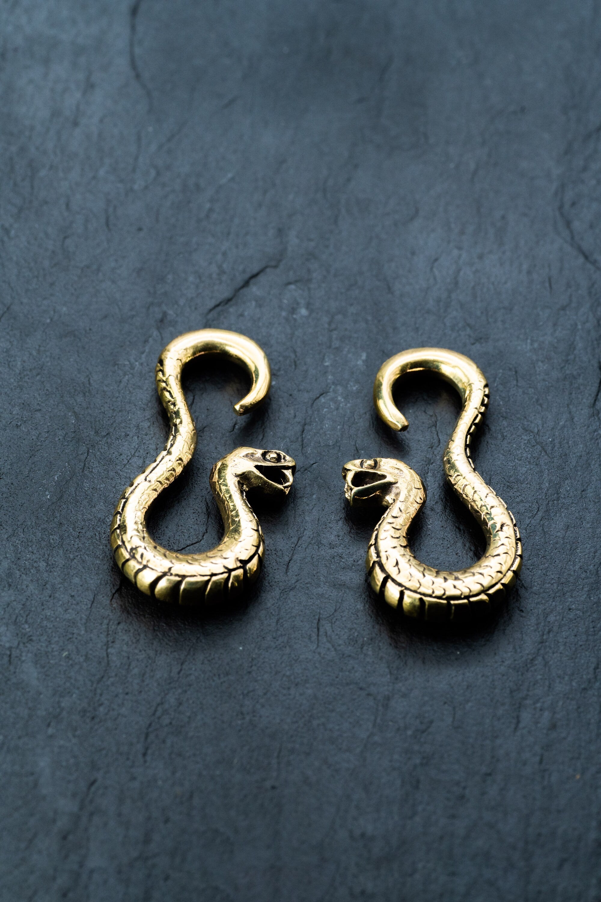 Two intricately designed Rattlesnake Gold Ear Weights from Kaya Movement lie on a dark, textured surface. The brass snakes are positioned symmetrically, with their heads facing each other, displaying detailed scales and open mouths. These ear weights are reminiscent of antique jewelry and perfect for stretched lobes.