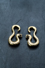 Two intricately designed Rattlesnake Gold Ear Weights from Kaya Movement lie on a dark, textured surface. The brass snakes are positioned symmetrically, with their heads facing each other, displaying detailed scales and open mouths. These ear weights are reminiscent of antique jewelry and perfect for stretched lobes.