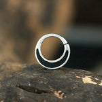 A close-up image of a Septum in 925 Silver with Black Mother Pearl Shell by Kaya Movement, partially black on one side, resting on a wooden surface. The background is blurred, highlighting the detailed design of the silver septum ring and its subtle black mother pearl shell accents.