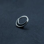 A close-up image of a small, elegant Kaya Movement Septum Ring in 925 Silver with Black Mother Pearl Shell placed on a smooth, dark surface. The earring is slightly open, showcasing its sleek and simple round design, perfect as septum jewelry.