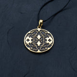 A Shipibo pendant from Kaya Movement, featuring intricate maze-like patterns and oxidized details, hangs from a black cord necklace on a dark, textured surface. The round, yellow brass pendant closely resembles Shipibo Icaros symbols, creating a sophisticated and detailed appearance with geometric shapes and lines.