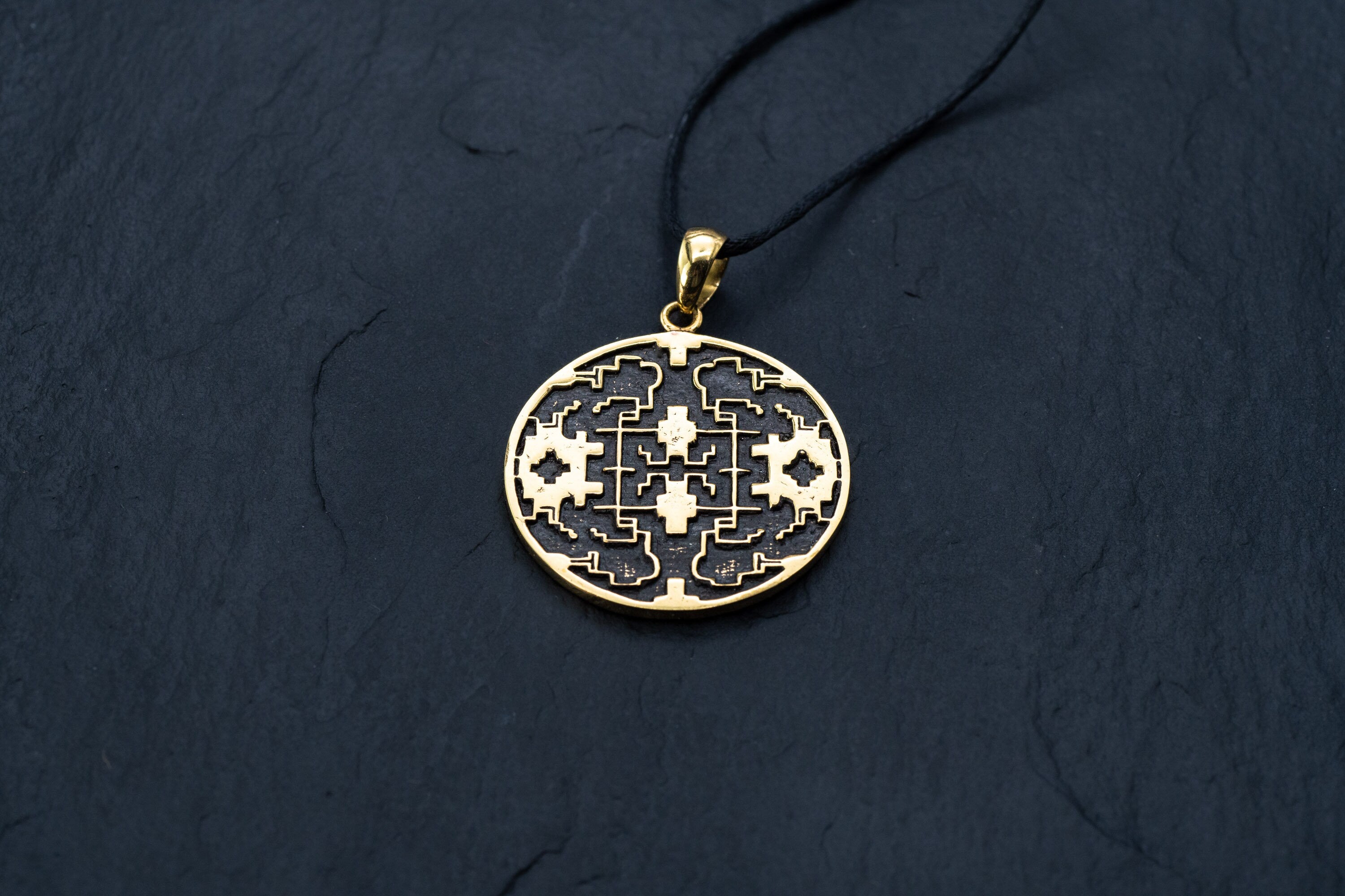 A Shipibo pendant from Kaya Movement, featuring intricate maze-like patterns and oxidized details, hangs from a black cord necklace on a dark, textured surface. The round, yellow brass pendant closely resembles Shipibo Icaros symbols, creating a sophisticated and detailed appearance with geometric shapes and lines.