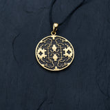 A close-up of the Kaya Movement Shipibo pendant from their Trippy Jewelry collection, crafted in yellow brass with intricate geometric patterns and oxidized details. The pendant hangs from a thin, black cord and is set against a dark, textured background. The design on the pendant features symmetrical and maze-like elements.