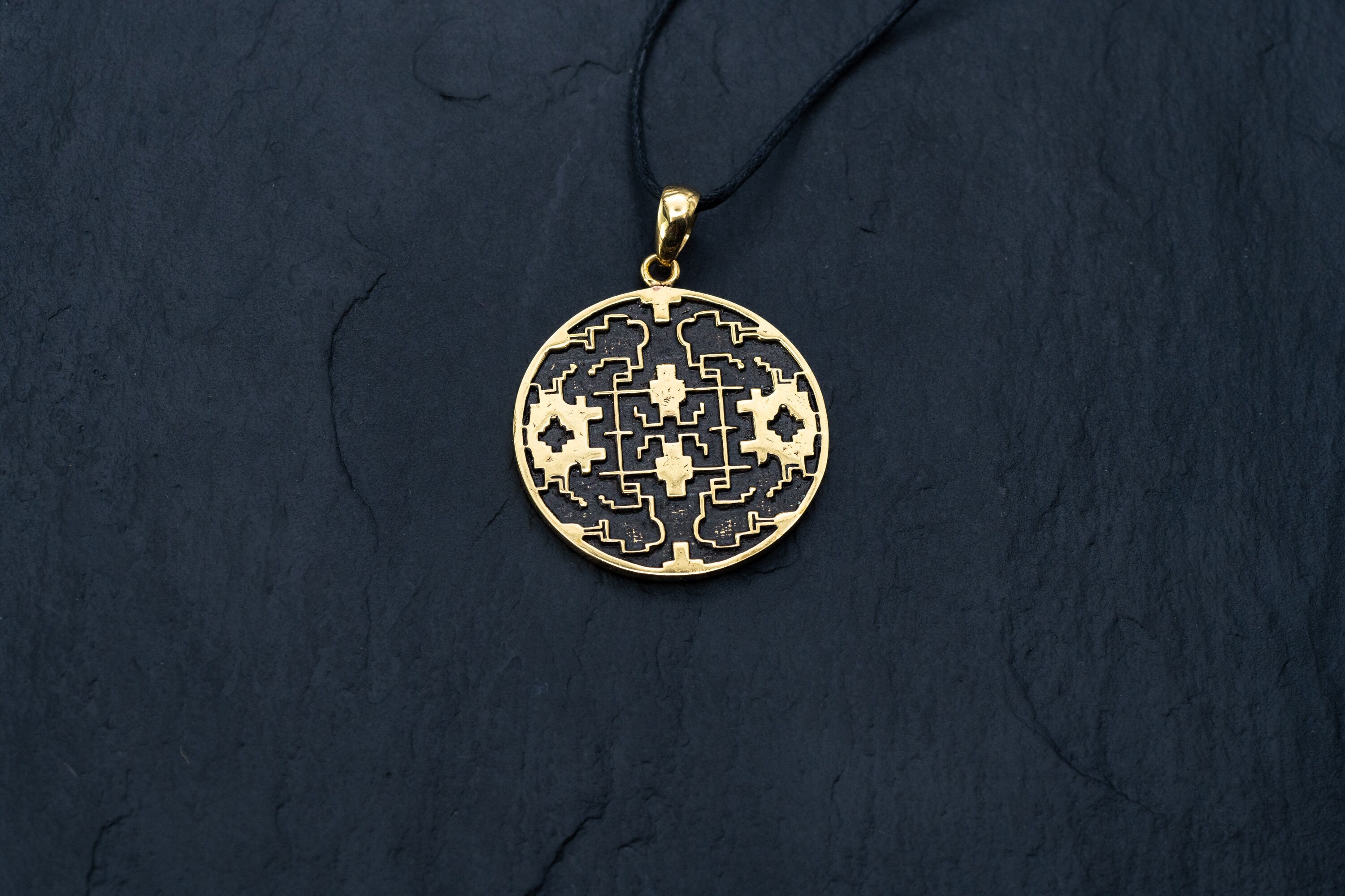 A close-up of the Kaya Movement Shipibo pendant from their Trippy Jewelry collection, crafted in yellow brass with intricate geometric patterns and oxidized details. The pendant hangs from a thin, black cord and is set against a dark, textured background. The design on the pendant features symmetrical and maze-like elements.