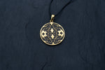 A close-up of the Kaya Movement Shipibo pendant from their Trippy Jewelry collection, crafted in yellow brass with intricate geometric patterns and oxidized details. The pendant hangs from a thin, black cord and is set against a dark, textured background. The design on the pendant features symmetrical and maze-like elements.