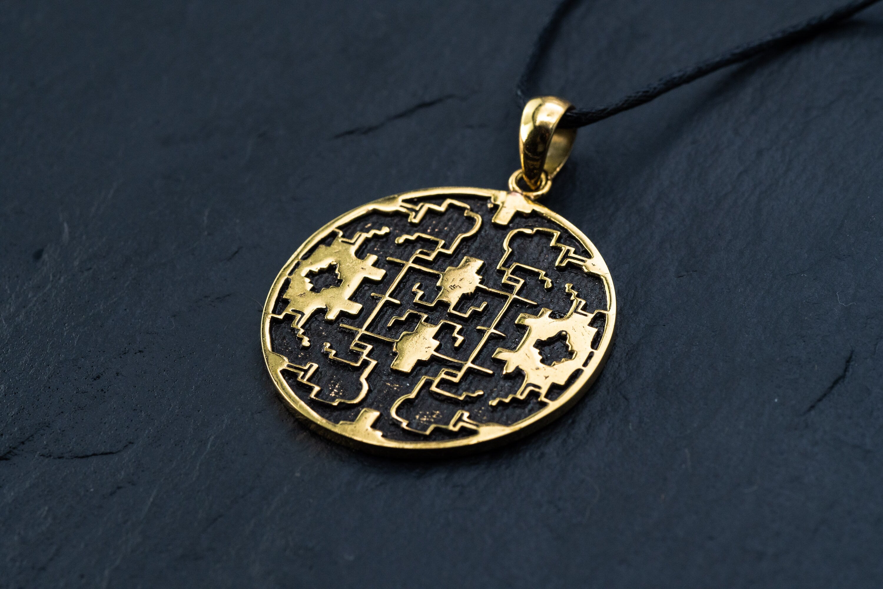 The Kaya Movement Shipibo pendant, known as Trippy jewelry, is a yellow brass shamanic pendant featuring intricate geometric and floral cutout designs. Attached to a black cord necklace, this psychedelic pendant showcases symmetrical patterns and oxidized details that shine against a dark background, highlighting its detailed craftsmanship.