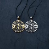 Two round Kaya Movement Shipibo Icaro pendants with intricate patterns featuring skull designs. One pendant is yellow brass with a black background and oxidized details, while the other is silver with a black background. Both are attached to black cords and set against a dark, textured surface.