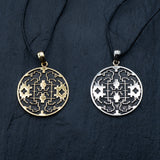 Two circular Shipibo pendants from Kaya Movement, featuring intricate geometric patterns, hang on black cords against a dark background. The left pendant, crafted from yellow brass, showcases oxidized details, while the right one has a silver-toned finish. The symmetrical designs on both shamanic pendants display lines and shapes that contribute to their complex and ornate look.