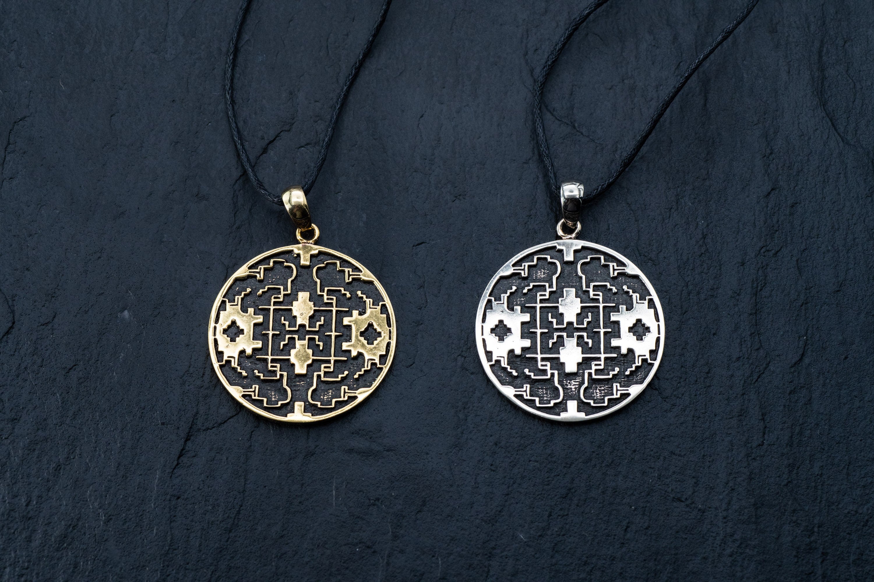 Two circular Shipibo pendants from Kaya Movement, featuring intricate geometric patterns, hang on black cords against a dark background. The left pendant, crafted from yellow brass, showcases oxidized details, while the right one has a silver-toned finish. The symmetrical designs on both shamanic pendants display lines and shapes that contribute to their complex and ornate look.