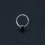 A minimalist silver ring featuring a single small black onyx gemstone centered at the front is set against a dark, textured background. The design, reminiscent of Kaya Movement's Black Onyx Silver Septum Jewelry – Seamless Nose Ring (8mm) for helix and daith piercings, is simple and elegant.