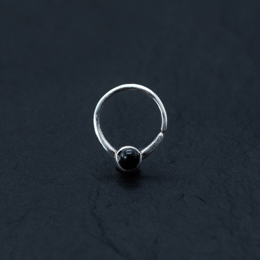 A minimalist silver ring featuring a single small black onyx gemstone centered at the front is set against a dark, textured background. The design, reminiscent of Kaya Movement's Black Onyx Silver Septum Jewelry – Seamless Nose Ring (8mm) for helix and daith piercings, is simple and elegant.