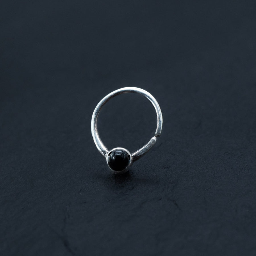 A simple yet sophisticated septum ring, featuring a smooth, round black onyx gemstone set in its center. This elegant piece, part of Kaya Movement's exclusive Silver 925 Jewelry collection, is displayed against a dark, textured background, highlighting its minimalist design and polished finish.