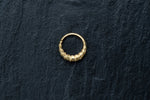 A small, intricately designed Biomechanical - 18k Gold Septum 10mm by Kaya Movement lies on a dark textured surface. The ring boasts a unique geometric pattern with raised square elements, giving it a distinct appearance reminiscent of futuristic gold-plated septum jewelry. The background contrasts sharply with the shiny gold, highlighting the ring's details.