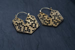 The Kaya Movement Oversized Hoop Honeycomb Hexagon Bee Gold Brass Earrings are displayed on a dark slate background. These closed hoop earrings feature a striking hexagonal shape with interconnected cells, evoking a beehive structure and embodying the essence of sacred geometry in cool boho hypercube jewelry.