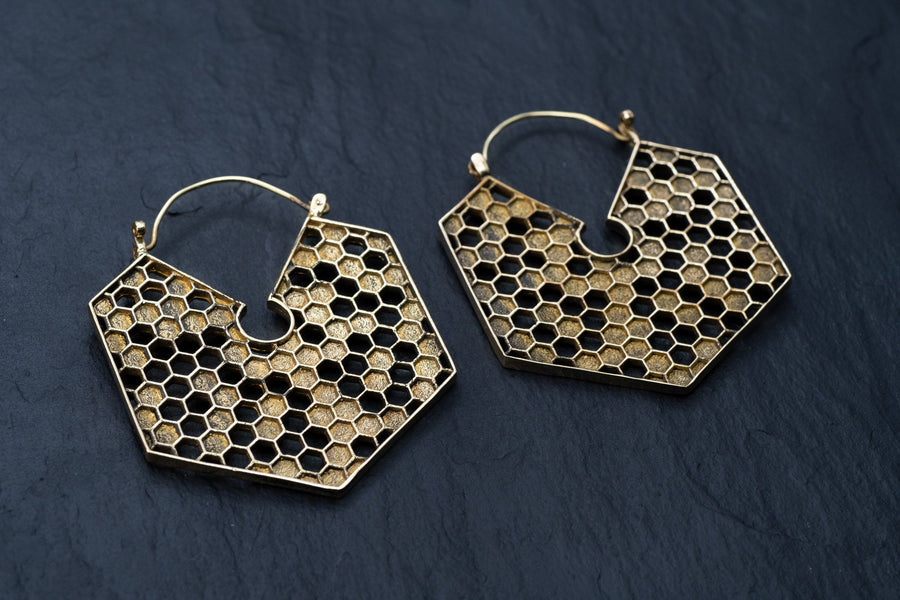 A pair of Kaya Movement's oversized hoop honeycomb hexagon bee gold brass earrings lies on a dark slate surface. These Sacred Geometry Cool Boho Hypercube Jewelry pieces feature a hollowed-out center and showcase an intricate and detailed design in a closed hoop style.