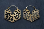 The Oversized Hoop Honeycomb Hexagon Bee Gold Brass Earrings from Kaya Movement, with their intricate honeycomb patterns, are placed on a dark, textured background. These geometric and stylish earrings exhibit sacred geometry and boho hypercube design, perfect for any occasion.