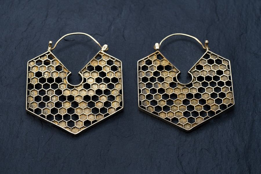 The Oversized Hoop Honeycomb Hexagon Bee Gold Brass Earrings from Kaya Movement, with their intricate honeycomb patterns, are placed on a dark, textured background. These geometric and stylish earrings exhibit sacred geometry and boho hypercube design, perfect for any occasion.