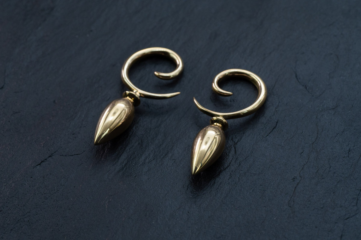A pair of Drop Ear Hangers by Kaya Movement, featuring shiny gold spirals with teardrop-shaped ends, placed on a dark slate background. These 4g Spiral Gauges curve inward to create an elegant and modern design. Hand-carved and perfect for body modification or stretched ears, these exquisite Gold Hangers add a touch of sophistication to any look.