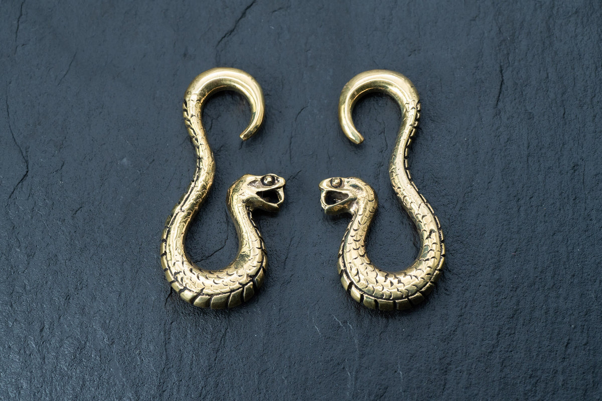A pair of Kaya Movement's Rattlesnake Gold Ear Weights, intricately designed with detailed scales and open-mouthed heads, are displayed side by side on a dark textured background. These snake-shaped brass ear weights curve elegantly, resembling the shape of question marks.
