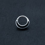A close-up image of a small, circular Septum in 925 Silver with Black Mother Pearl Shell by Kaya Movement, placed against a dark, textured background. The earring has a simple, minimalist design with a slight curve, reminiscent of sleek septum jewelry.