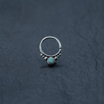 Introducing the Opal Septum Ring Silver by Kaya Movement: a seamless teardrop-shaped septum ring crafted from sterling silver, featuring a small, round opal stone at its center and adorned with a cluster of tiny silver beads. This 18g, 8mm nose ring is set against a dark, textured background that highlights its intricate design and exceptional craftsmanship.