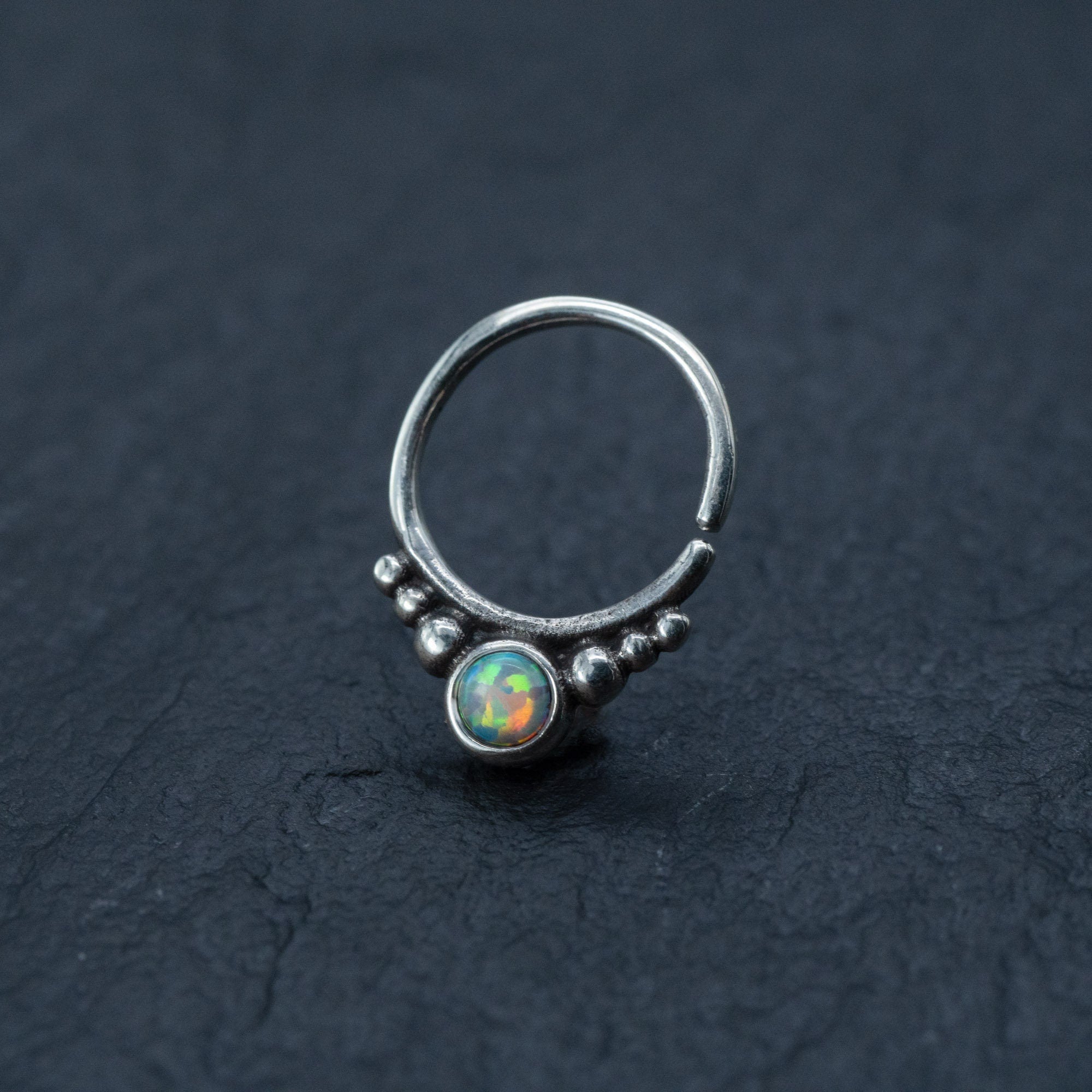 A close-up image of the Kaya Movement Opal Septum Ring Silver, featuring a translucent opal stone at its center in a teardrop design, surrounded by small silver beads. This seamless 18g septum jewelry is positioned on a dark, textured surface, accentuating the intricate details and colorful reflections of the 8mm opal.