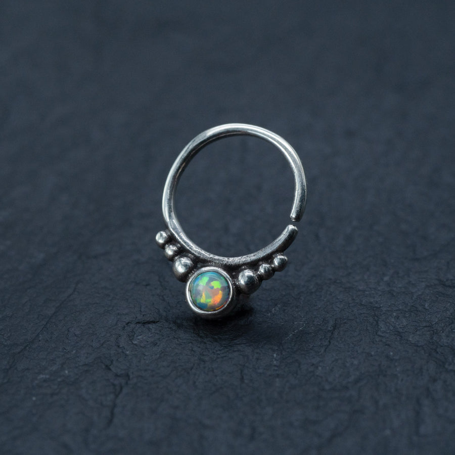 A close-up image of the Kaya Movement Opal Septum Ring Silver, featuring a translucent opal stone at its center in a teardrop design, surrounded by small silver beads. This seamless 18g septum jewelry is positioned on a dark, textured surface, accentuating the intricate details and colorful reflections of the 8mm opal.