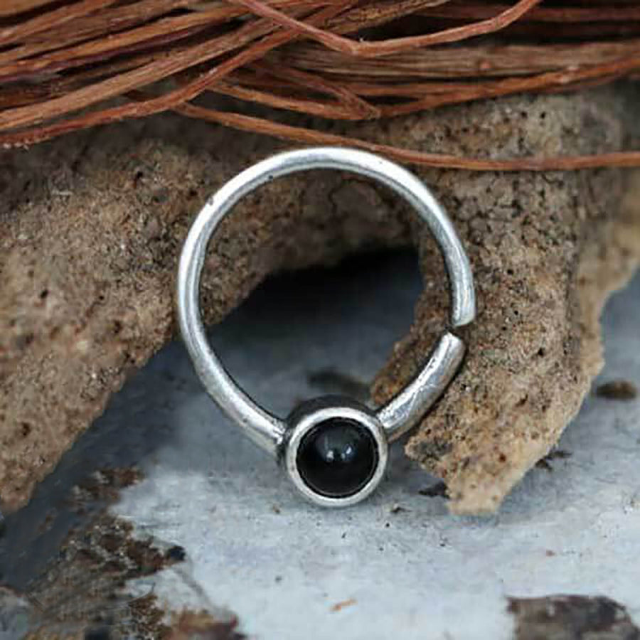 A close-up of the Kaya Movement Black Onyx Silver Septum Jewelry, showcasing an 8mm seamless nose ring adorned with a circular black onyx gemstone at the opening. The piece is displayed against a textured, earthy background accented with hints of dried plant material.
