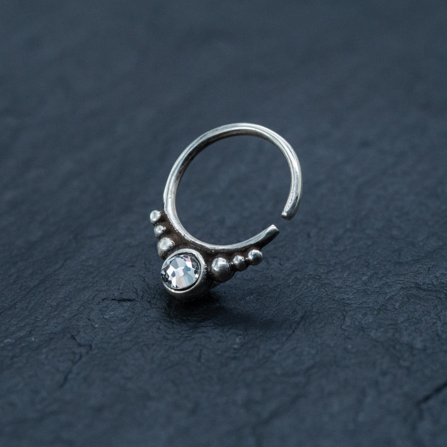 A close-up of the Kaya Movement Zircon Silver Nose Septum Ring Piercing resting on a textured dark surface. The Daith jewelry features a clear, round gemstone at its center and a patterned design with small silver beads on either side of the stone. This 8mm teardrop septum ring embodies both gothic charm and cuteness.