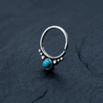 A silver septum ring from Kaya Movement is placed on a dark textured background. The ring features a small, round turquoise gemstone set in the center with tiny silver beads arranged around it in an ornate design. Made from 925 silver, this Bohemian-style jewelry has an open end, indicating it is adjustable.