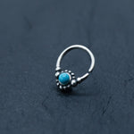 A small Kaya Movement Silver septum ring with an open end lies on a dark textured surface. The exquisite ring, part of their Bohemian jewelry collection, features a central turquoise gemstone surrounded by delicate silver beads.