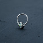 A Kaya Movement Silver septum ring with a small, round, iridescent opal gemstone in the center, surrounded by tiny silver beads, is placed on a dark gray surface. The elegant curved ring is open, highlighting its refined design.