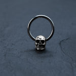Displayed on a dark, textured surface, the Skull Ball Closure Rings piercing from Kaya Movement features a small, metallic hoop made from Stainless Steel 316L. Attached at the bottom is a detailed skull charm with hollow eye sockets, a nose cavity, and intricately carved teeth that give it a realistic appearance, ideal for piercings. These silver skull earrings are perfect for those who love Punk Goth hoop earrings with charm and come in sizes 18g, 16g, and 14g.