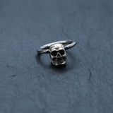 A silver Skull Ball Closure Ring, branded by Kaya Movement and crafted from high-quality Stainless Steel 316L, lies on a dark, textured surface. The punk goth hoop earring's simple band contrasts with the intricately detailed skull charm at its focal point, creating a striking visual effect ideal for piercings. Available in various gauge sizes—18g, 16g, and 14g—this piece is perfect for adding an edgy touch to your look.