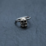A silver Skull Ball Closure Ring, branded by Kaya Movement and crafted from high-quality Stainless Steel 316L, lies on a dark, textured surface. The punk goth hoop earring's simple band contrasts with the intricately detailed skull charm at its focal point, creating a striking visual effect ideal for piercings. Available in various gauge sizes—18g, 16g, and 14g—this piece is perfect for adding an edgy touch to your look.