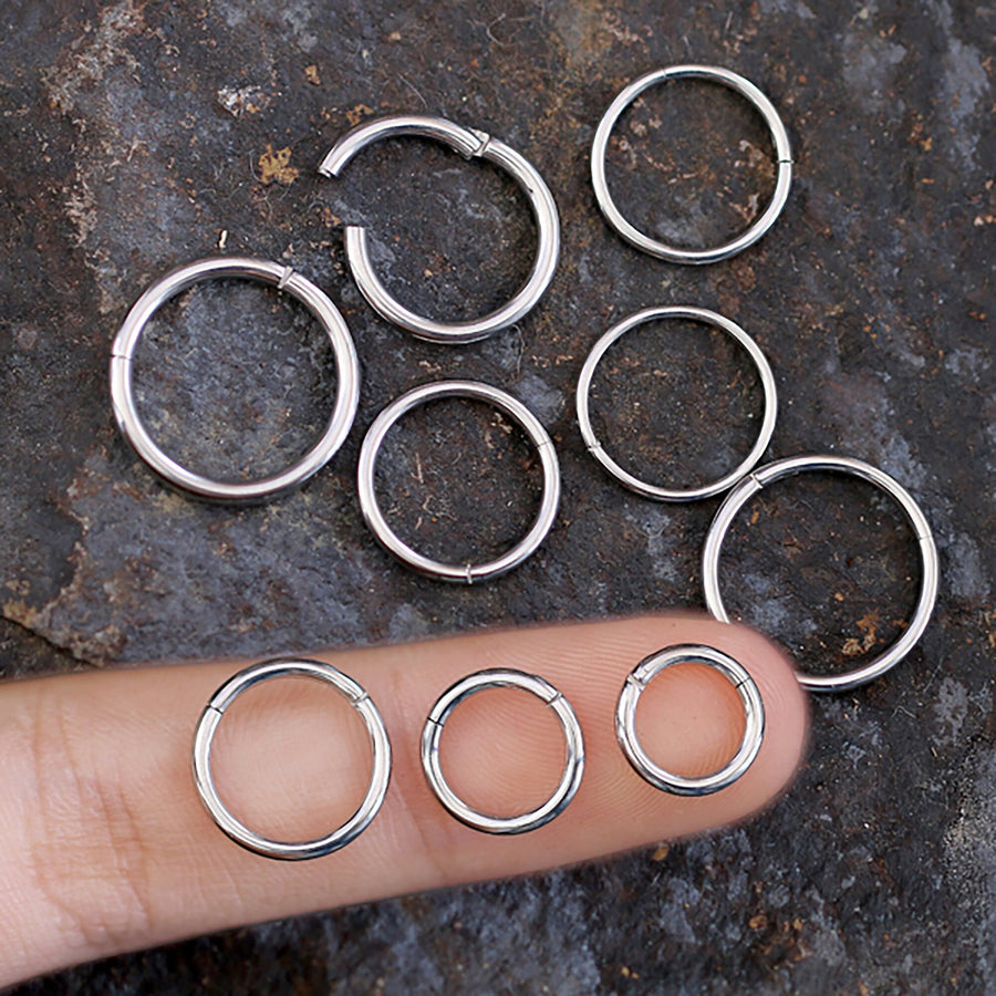 A range of silver-colored circular earrings, including tiny studs and Minimal Silver Hinged Clicker septums from Kaya Movement, are displayed on a rough, textured surface. Four earrings are arranged in a semi-circle. A person's finger is pointing to and holding two differently sized 316L Surgical Steel Silver septum clicker rings for comparison.