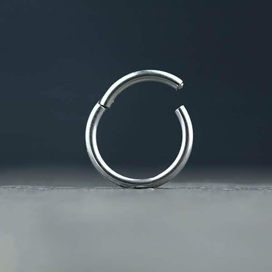 A minimalist, partially closed silver septum clicker ring from Kaya Movement against a dark backdrop. Made from 316L surgical steel, the tiny hinged septum ring is open at one end, with a sleek and smooth finish, highlighting its contemporary design akin to the Silver Septum Clicker Ring 16g.