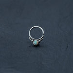 A delicate Opal Septum Ring Silver by Kaya Movement, featuring a small opal stone and accented with silver beads on either side, is placed on a dark, textured surface.